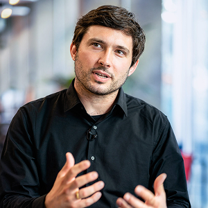 Photo rapprochée de Matthias Ott en train de parler. C'est un homme âgé d'une trentaine d'année aux cheveux bruns et courts. L'arrière de la photo est floutée, on ne voit que cet homme de manière nette